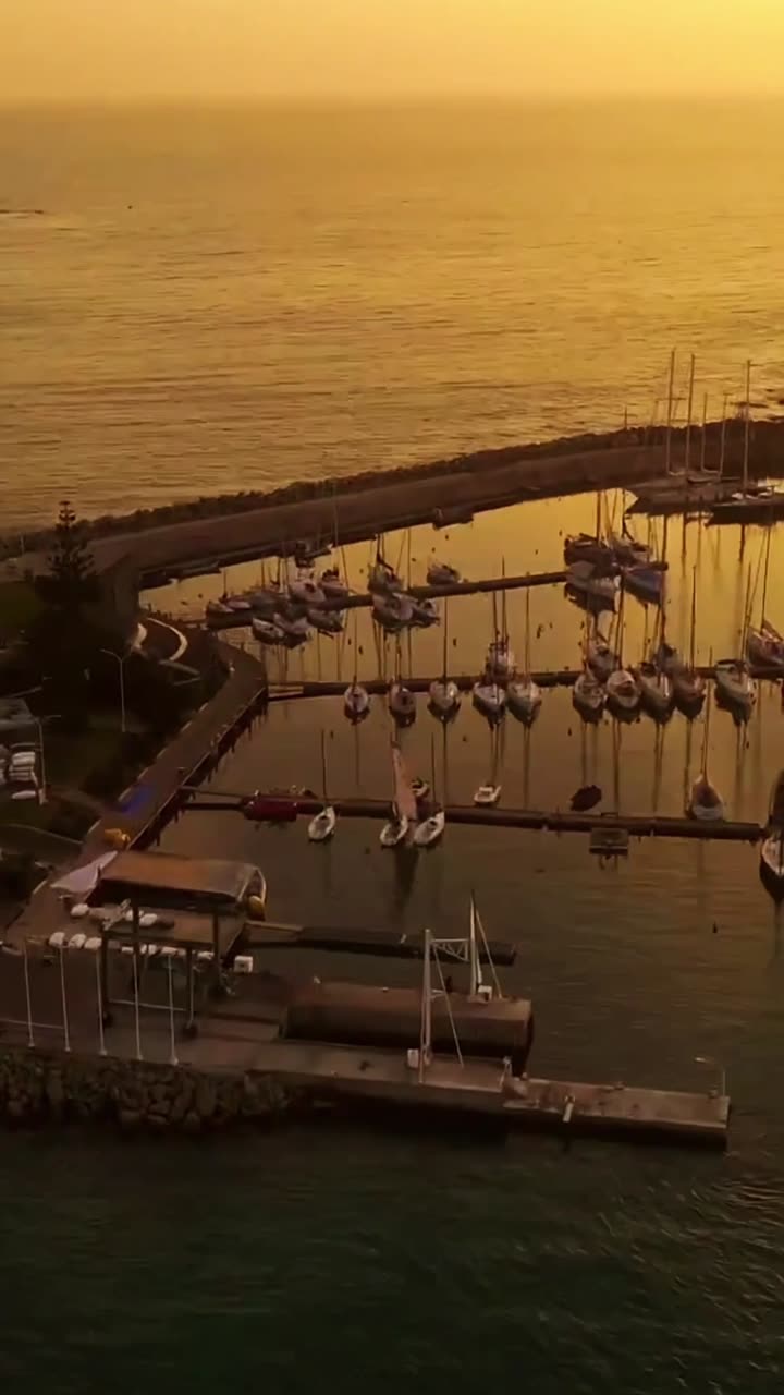 Toma aérea del puerto deportivo al atardecer, con veleros amarrados y el mar dorado al fondo